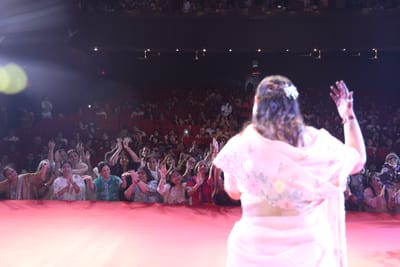 Anshu facing the packed hall from the stage with one hand raised, the balcony full above rows of red seats.