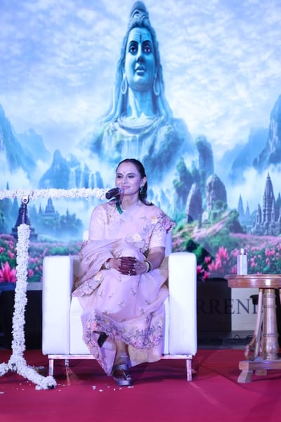 Anshu seated with a microphone on stage in front of a painted Himalayan backdrop.