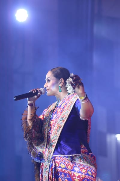 Anshu singing into a handheld microphone in a blue and red patola sari under a single spotlight.