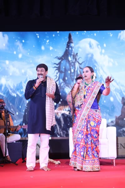 Anshu and a fellow singer sharing the stage, both on the microphone, against a painted backdrop.