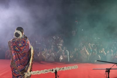 Anshu facing the audience across the red stage through green-lit haze, a jasmine garland strung along the microphone stand.