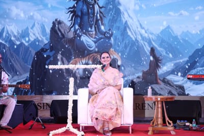 Anshu seated on stage with a jasmine-wrapped microphone stand, a painted Himalayan Shiva backdrop behind her.