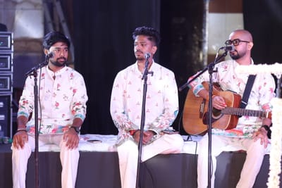 Three of the band seated along the stage riser at their microphones, one playing an acoustic guitar.