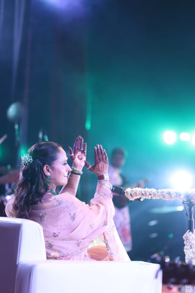 Anshu leaning forward with her hands open towards the hall, stage lights breaking green behind her.