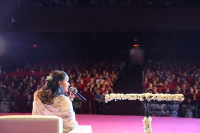 Anshu singing out to a full, darkened hall from her chair at the stage edge, the jasmine-wrapped microphone stand beside her.