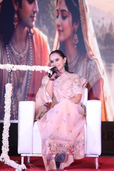 Anshu smiling into a handheld microphone from a white armchair, a large painted devotional mural behind her.