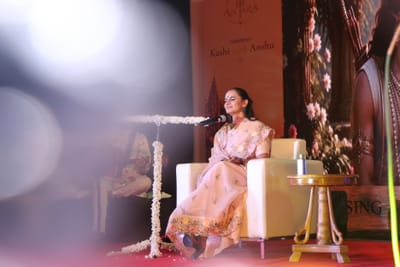 Anshu seated with her eyes closed mid-bhajan, the Antara and Kashi with Anshu banner behind the stage.