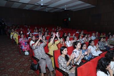 A wide view of the seated hall applauding, hands raised in namaste through the rows.