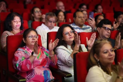 Two women in the audience clapping along, rows of red seats filled behind them.